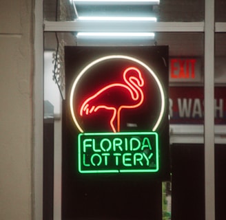 A neon sign depicting a flamingo above the words 'Florida Lottery' is prominently displayed. The sign is illuminated in bright red and green colors, and it is framed by glass panels. An 'EXIT' sign is visible in the background.