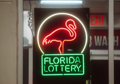 A neon sign depicting a flamingo above the words 'Florida Lottery' is prominently displayed. The sign is illuminated in bright red and green colors, and it is framed by glass panels. An 'EXIT' sign is visible in the background.