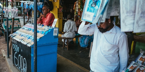 A street vendor stall displaying an array of lottery tickets clipped to a bright blue kiosk. A man in a red shirt leans on the counter, appearing thoughtful, while another man in a white shirt adjusts his hat and walks by. A variety of products hangs from strings above, and a colorful display fills the background with vibrant signs and packets. The scene suggests a busy, lively marketplace.